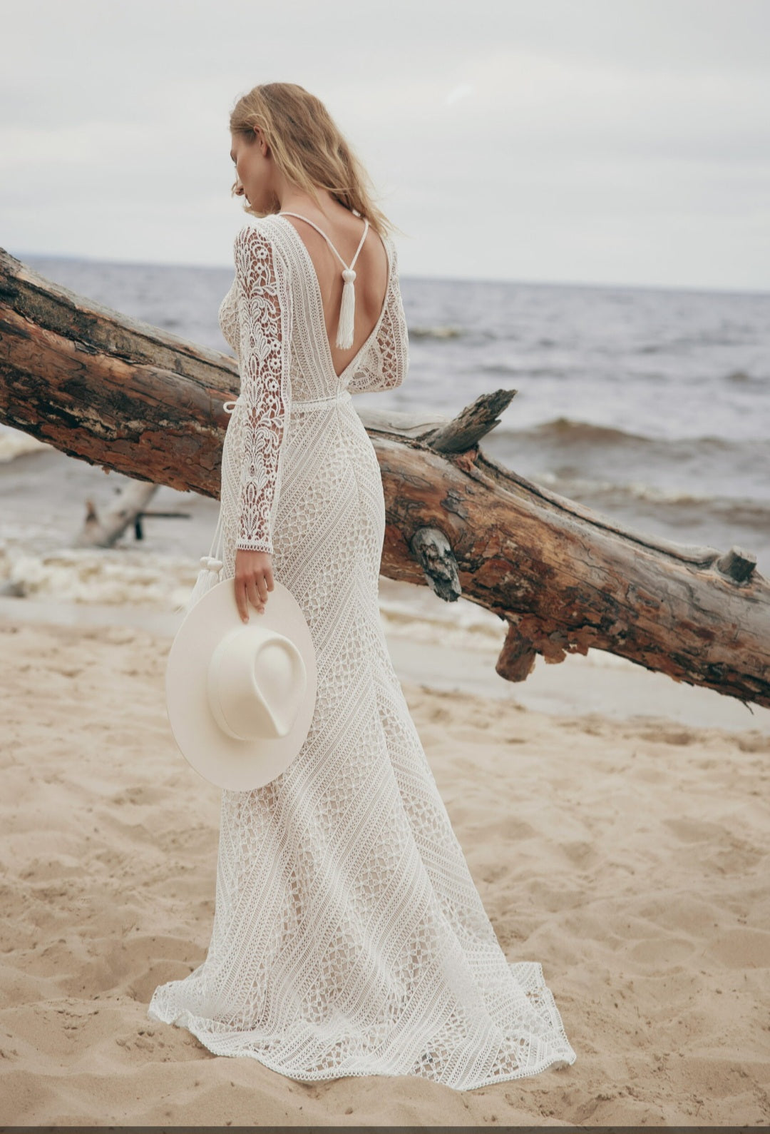 Woman in a white lace dress standing on a beach with a large log and ocean in the background