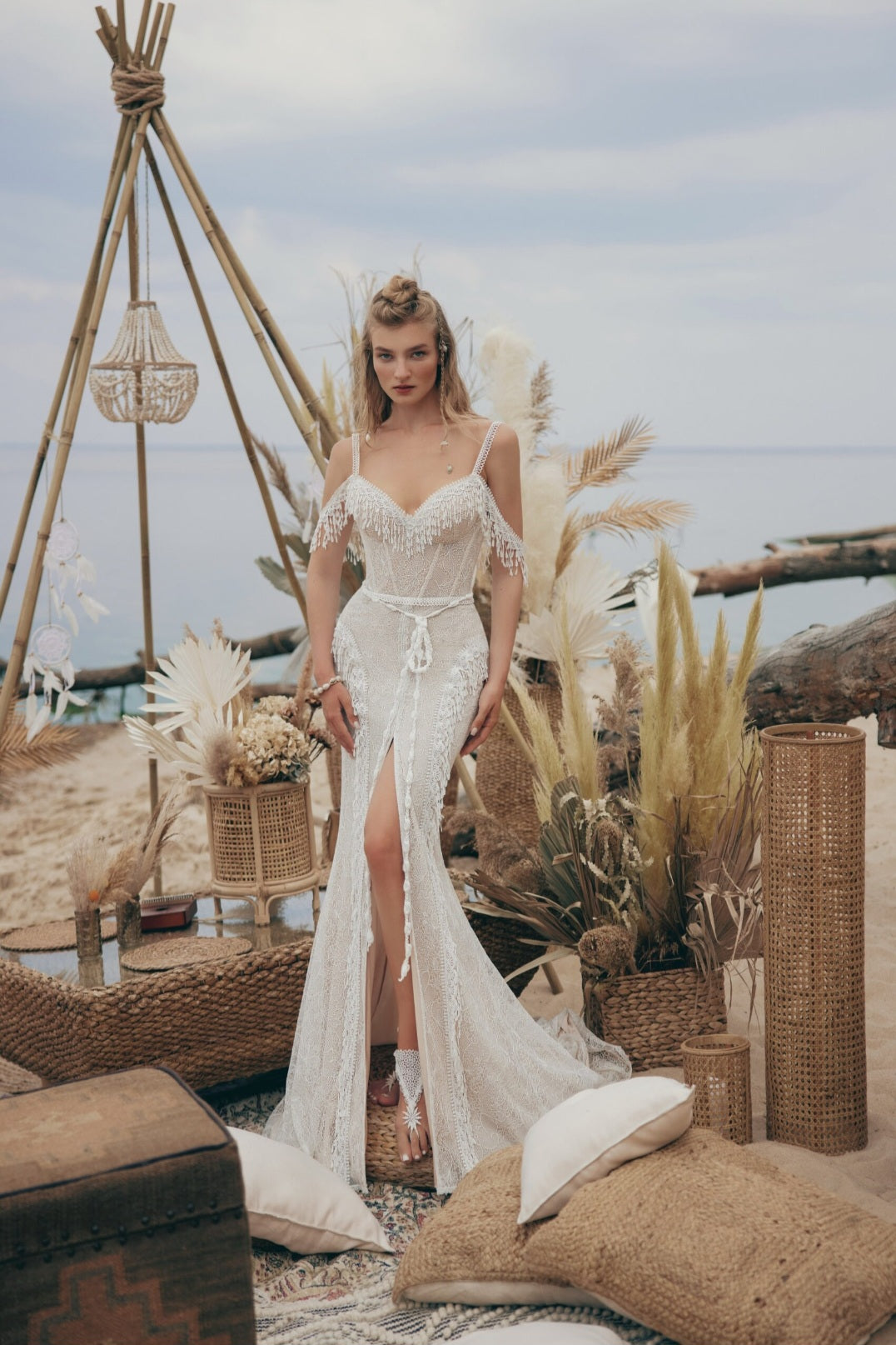 Woman in a white lace dress standing in a rustic outdoor setting with plants and a thatched roof.