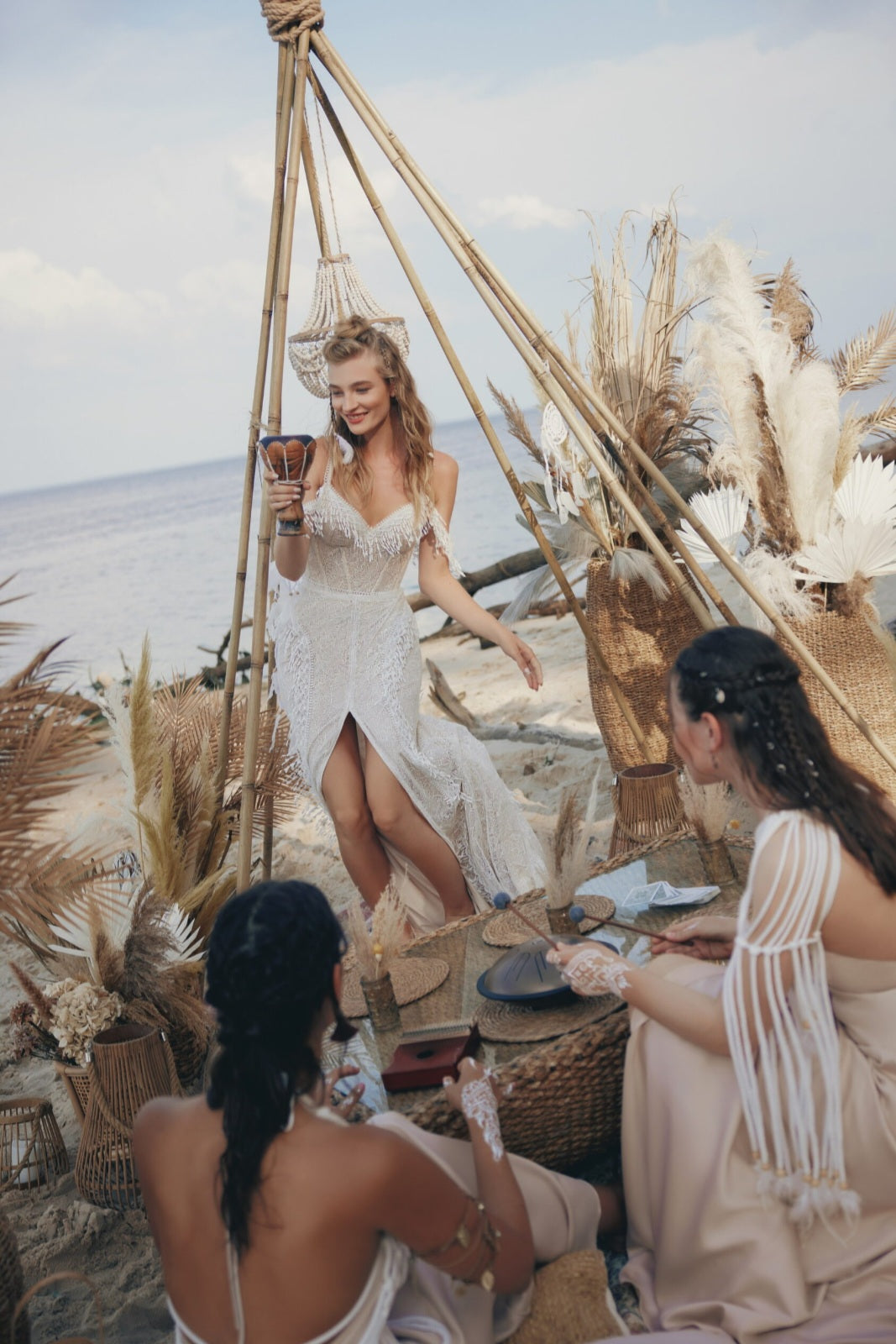 Woman in a white dress standing under a wooden canopy with two other women on a beach.