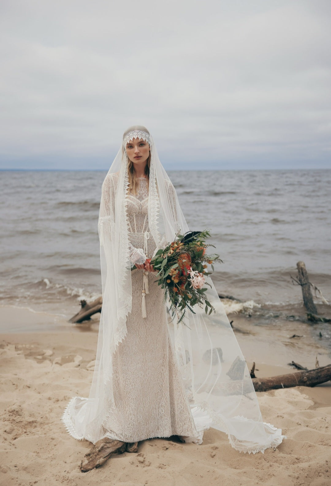 Woman in a wedding dress with a veil standing on a beach holding a bouquet.