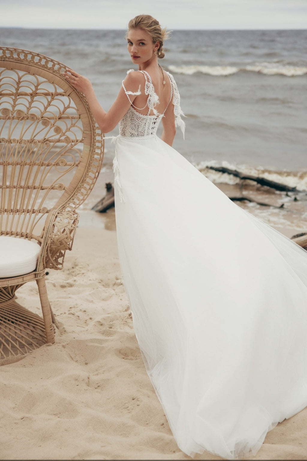 Woman in a white wedding dress standing on a beach with wicker chair and ocean view.