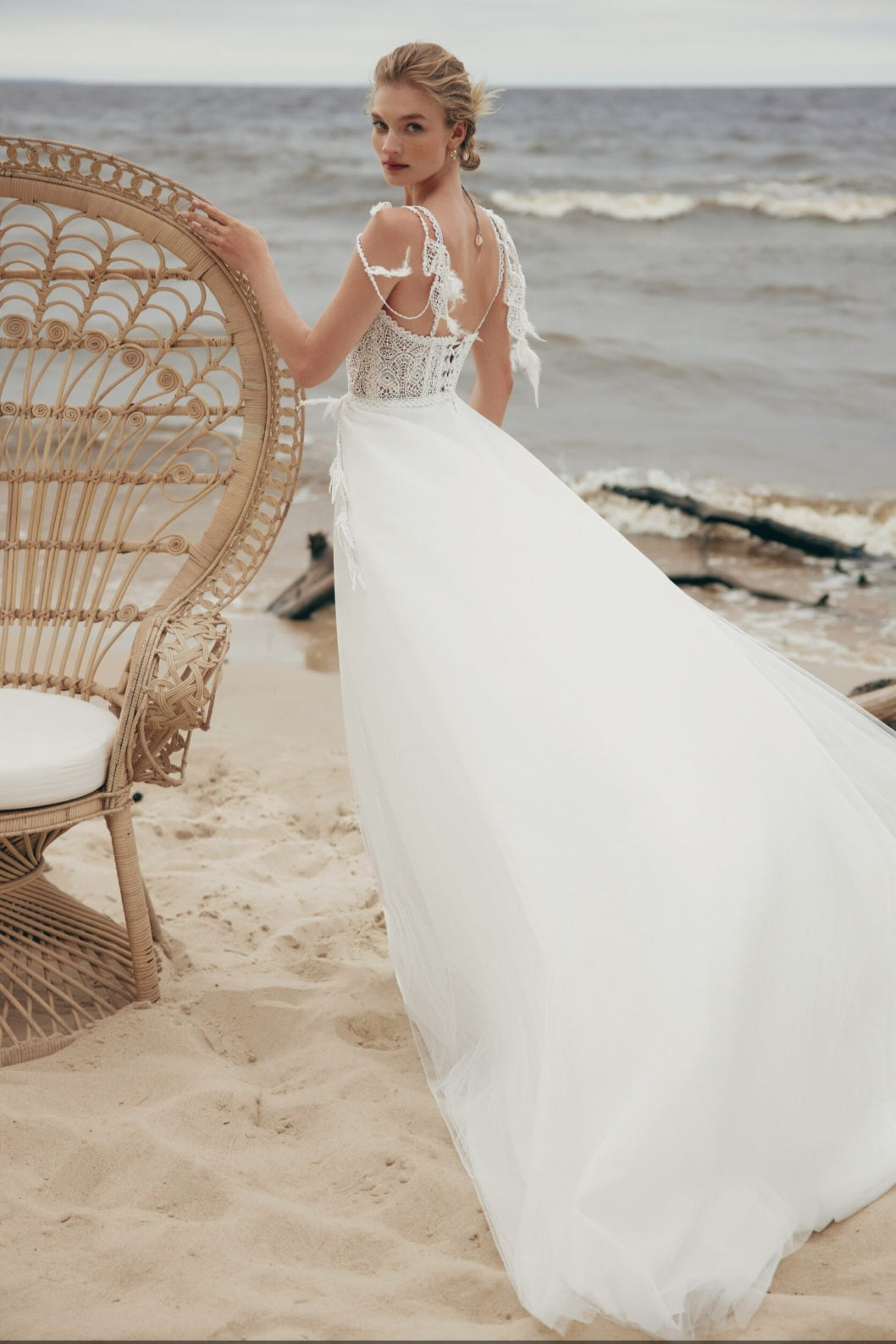 Woman in a white wedding dress standing on a beach with wicker chair and ocean view.