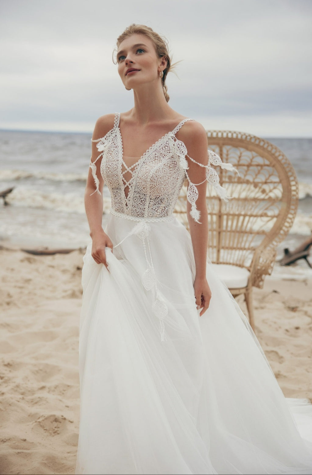 Woman in a white wedding dress standing on a beach with a wicker chair in the background.