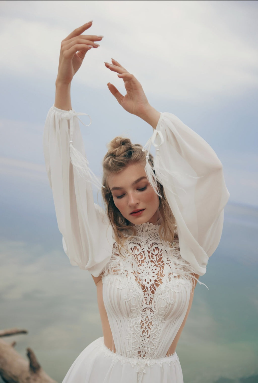 Woman in a white lace wedding dress with sheer sleeves against a blurred natural background