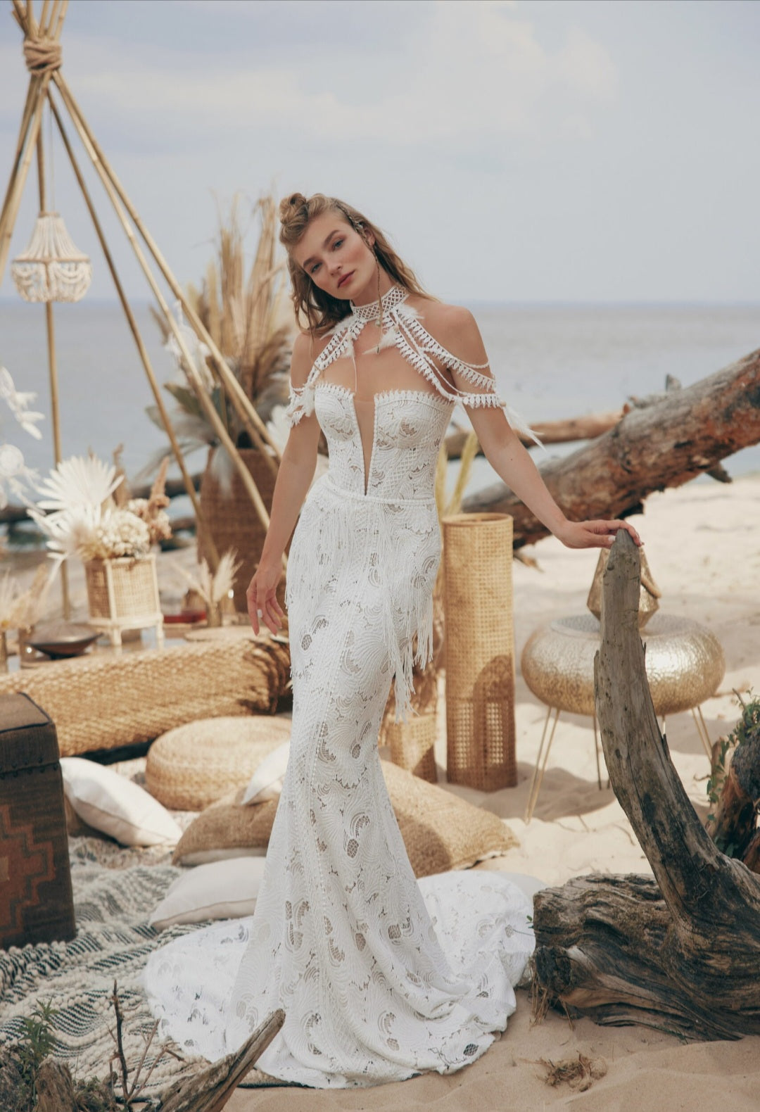 Woman in a white lace dress standing on a beach with driftwood and decorative elements.
