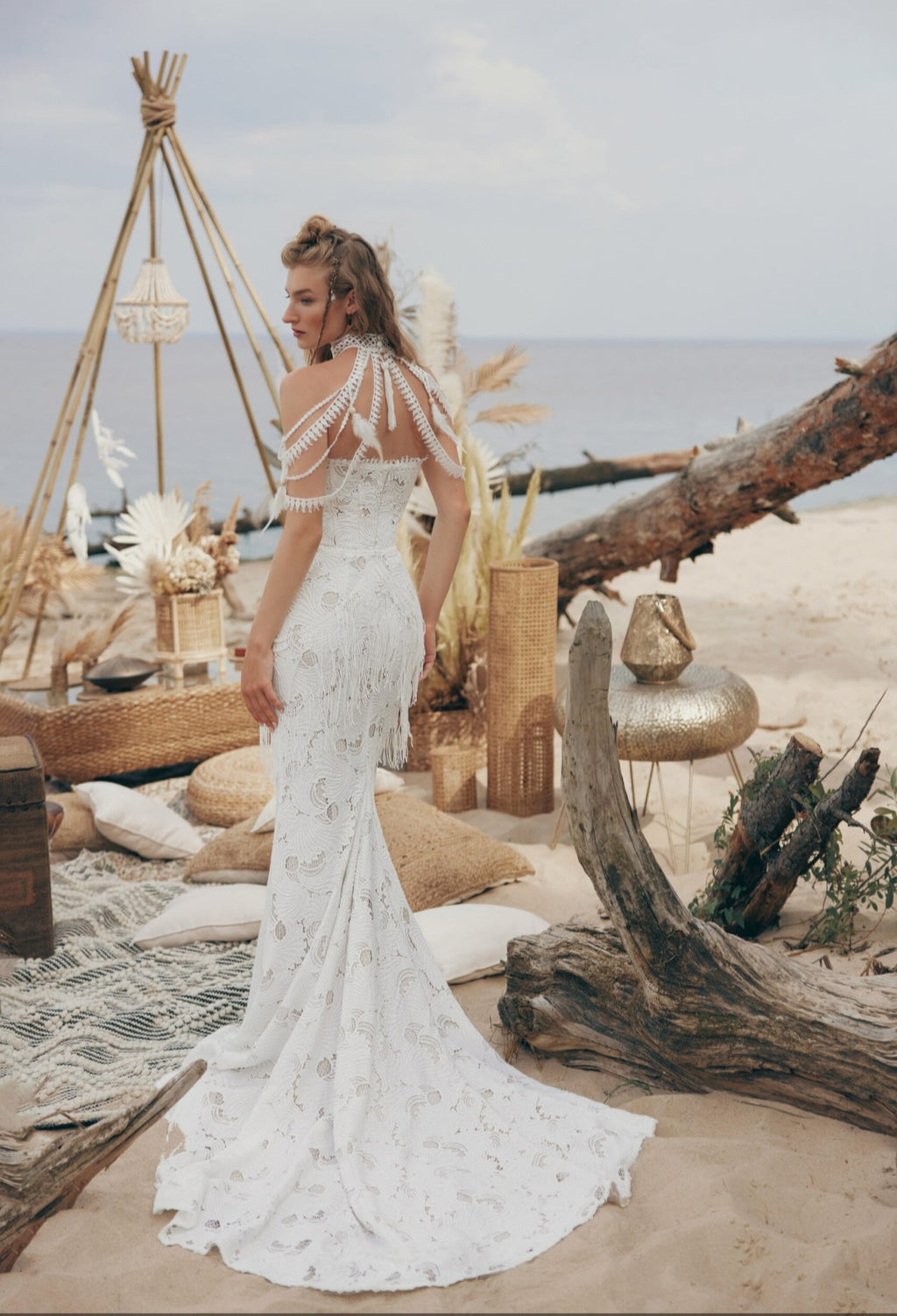 Woman in a white lace dress standing on a beach with driftwood and a teepee in the background