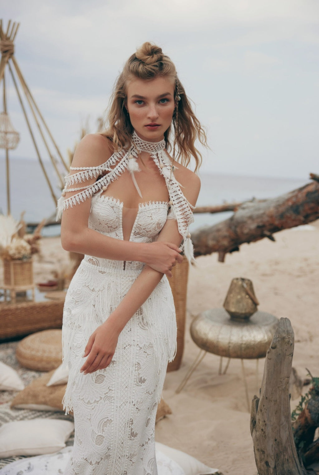 Woman in a white dress standing on a beach with driftwood and a tent-like structure in the background.