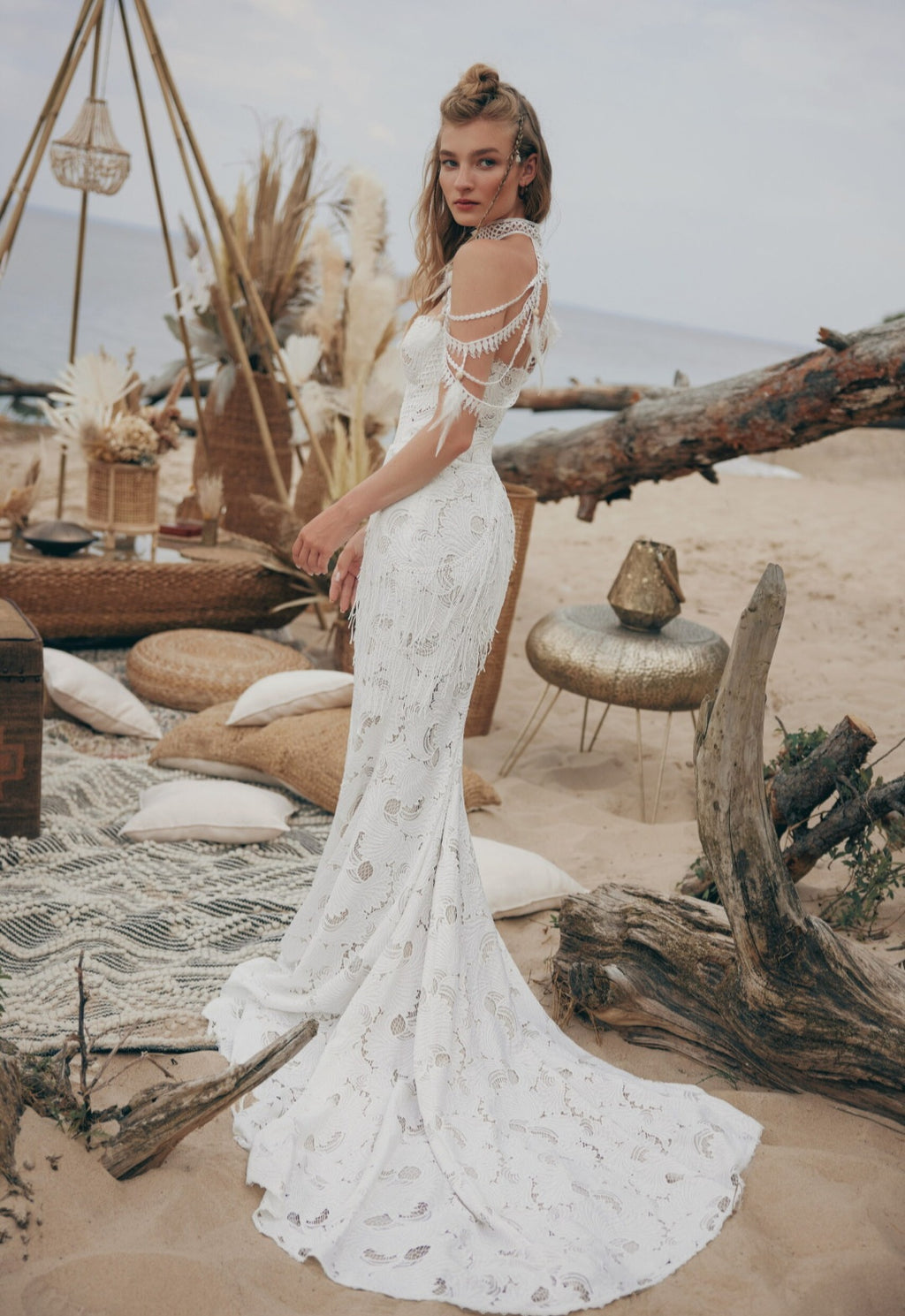 Woman in a white lace dress standing in a desert-like setting with driftwood and decorative elements.