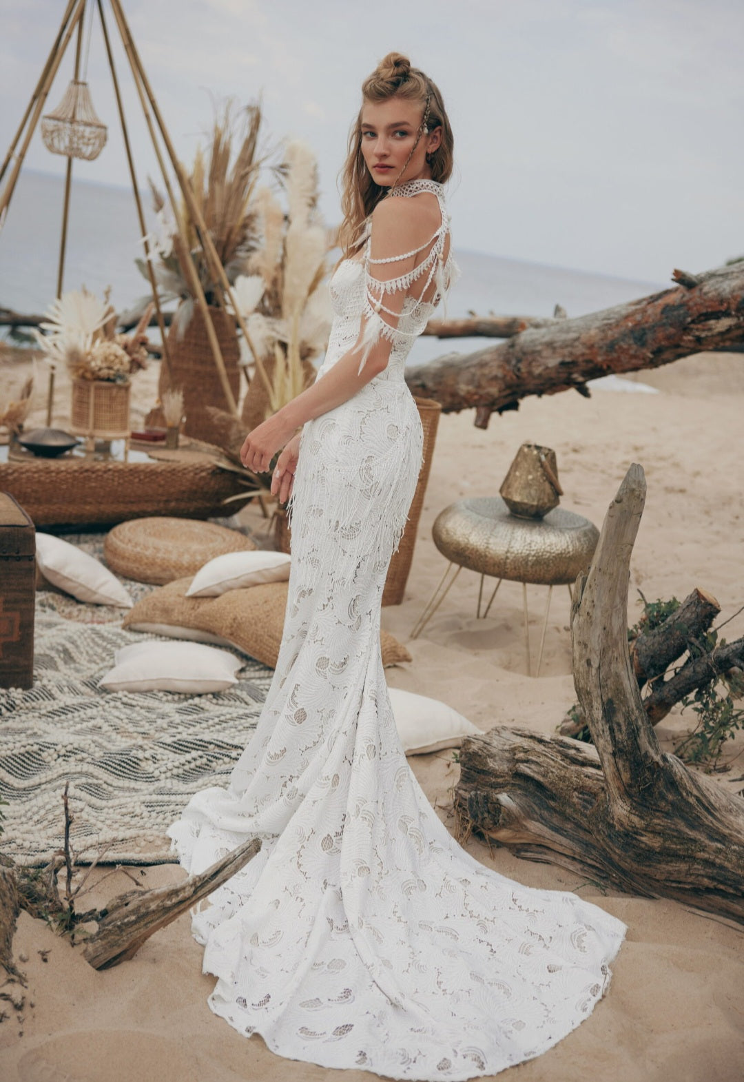 Woman in a white lace dress standing in a desert-like setting with driftwood and decorative elements.