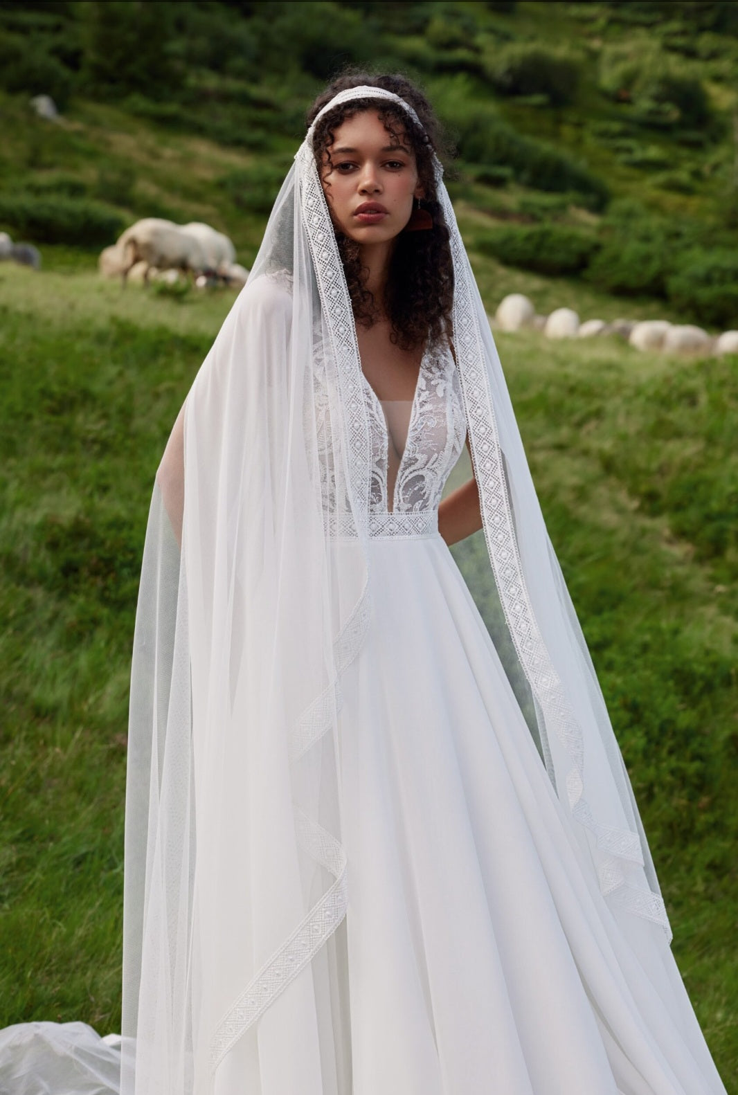 Woman in a white wedding dress with a long veil standing in a grassy field with sheep.