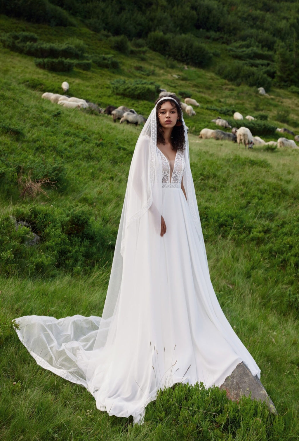 Woman in a white wedding dress with a hood standing in a grassy field with sheep.