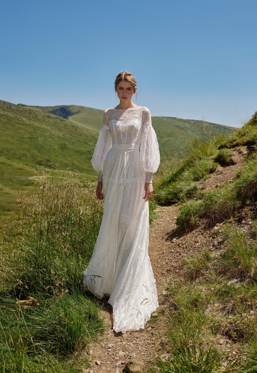 Woman in a white wedding dress standing on a mountain path with green hills and blue sky in the background.