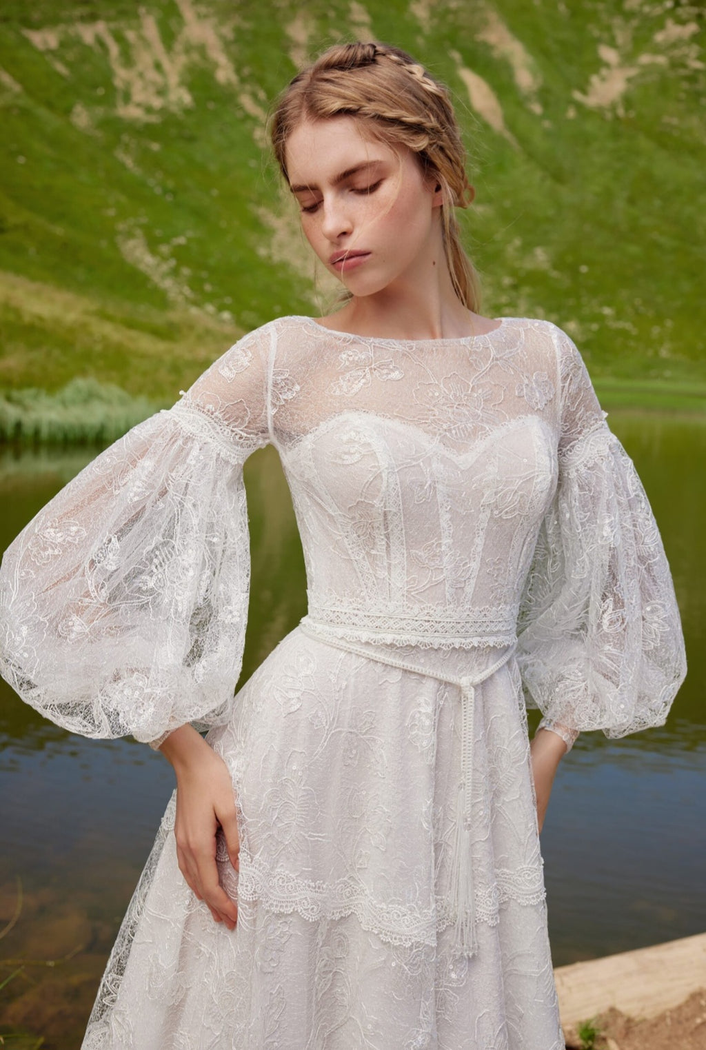 Woman wearing a white lace dress with long sleeves by a body of water.
