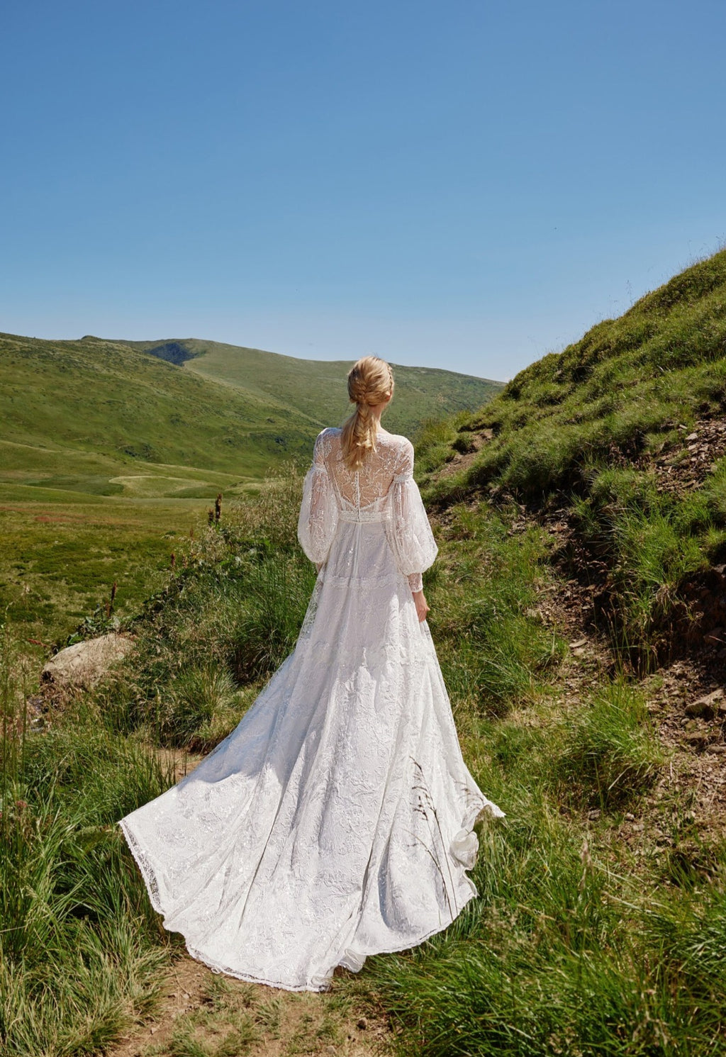 Woman in a white dress standing on a grassy hillside with a clear blue sky.