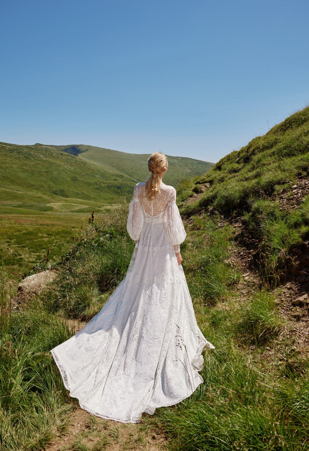 Woman in a white dress standing on a grassy hillside with a clear blue sky.