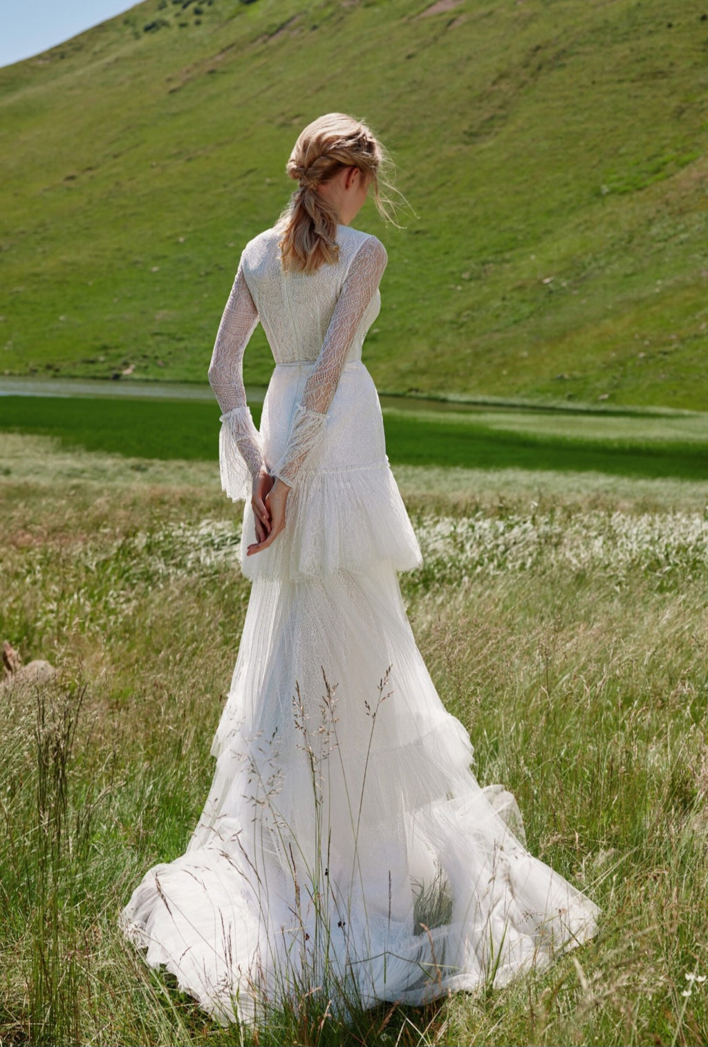 Woman in a white wedding dress standing in a grassy field with hills in the background