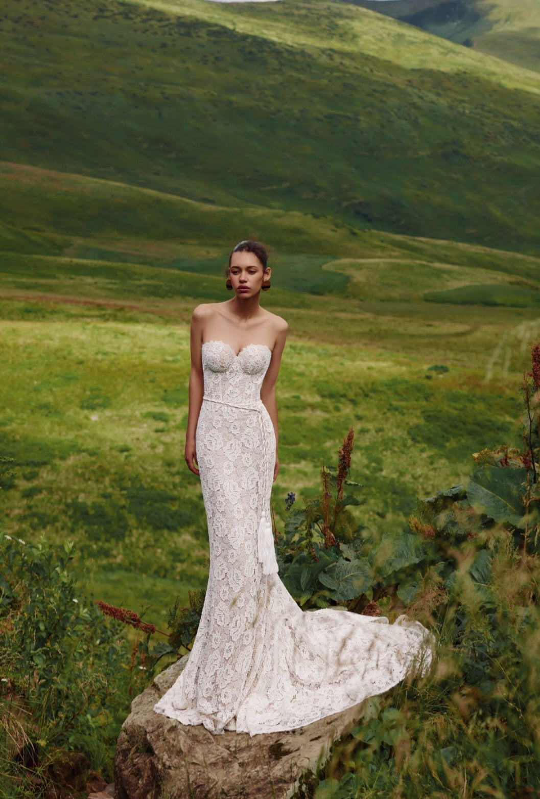 Woman in a white strapless gown standing on a rock with a scenic green landscape in the background