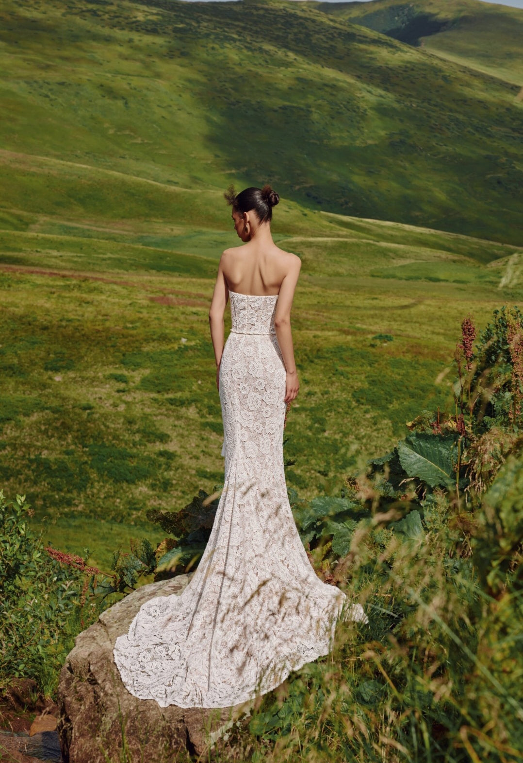 Woman in a white lace dress standing on a rocky outcrop with green hills in the background