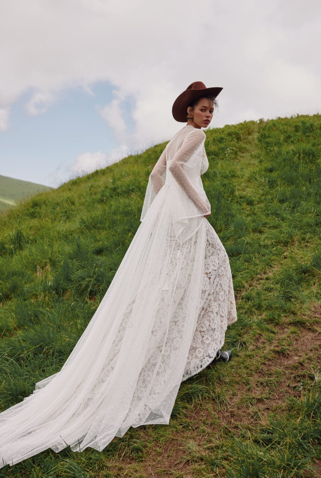 Woman in a white lace dress and brown hat standing on a grassy hillside.