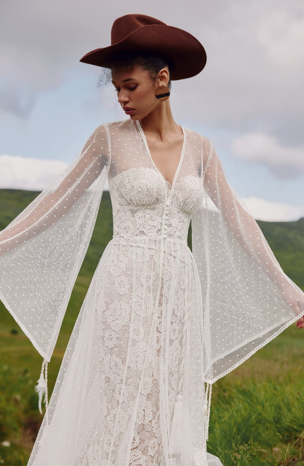 Woman in a lace dress with sheer sleeves and a wide-brimmed hat standing in a field.