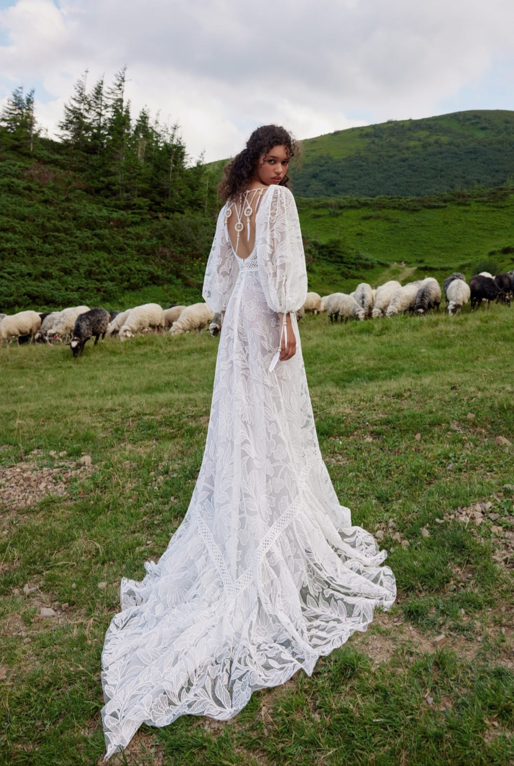 Woman in a white lace dress standing in a field with sheep and mountains in the background