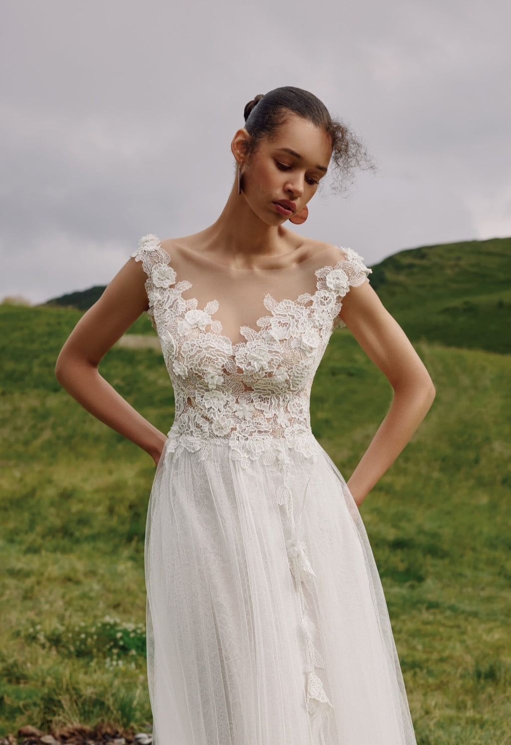 Woman in a white lace dress standing in a grassy field with hills in the background