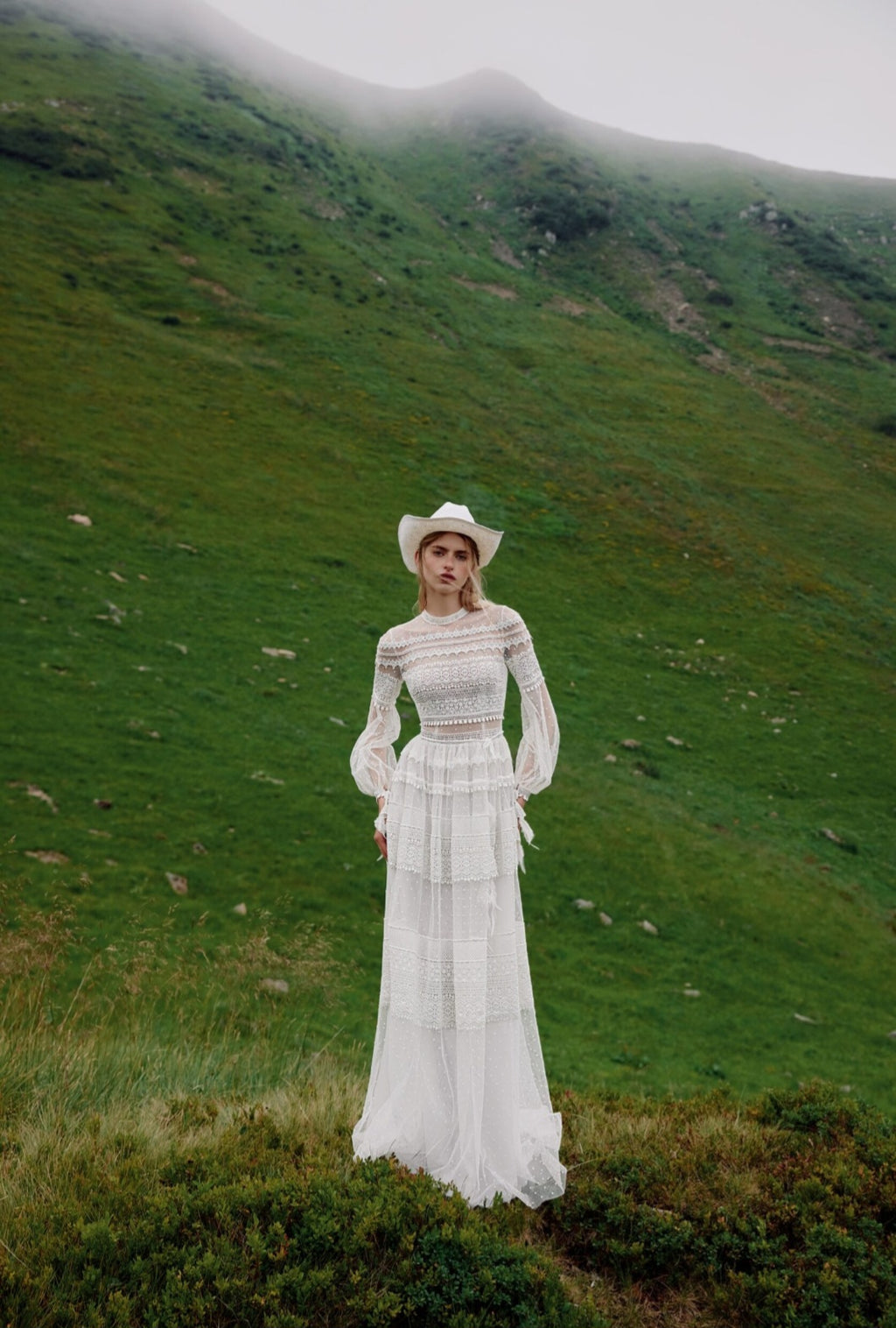 Woman in a white dress standing on a grassy hill with mountains in the background