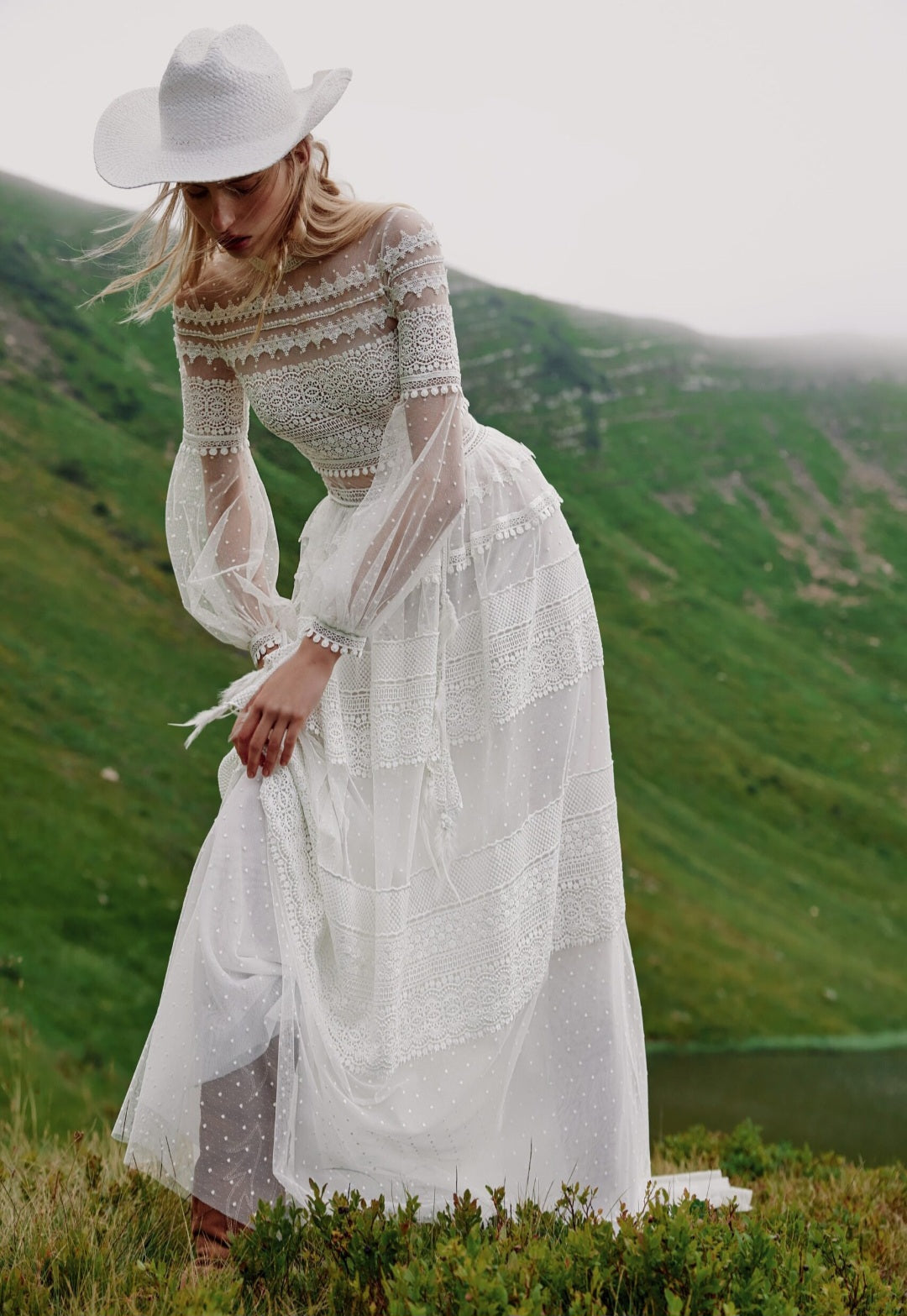 Woman in a white dress with long sleeves and a wide-brimmed hat standing in a grassy field with mountains in the background.