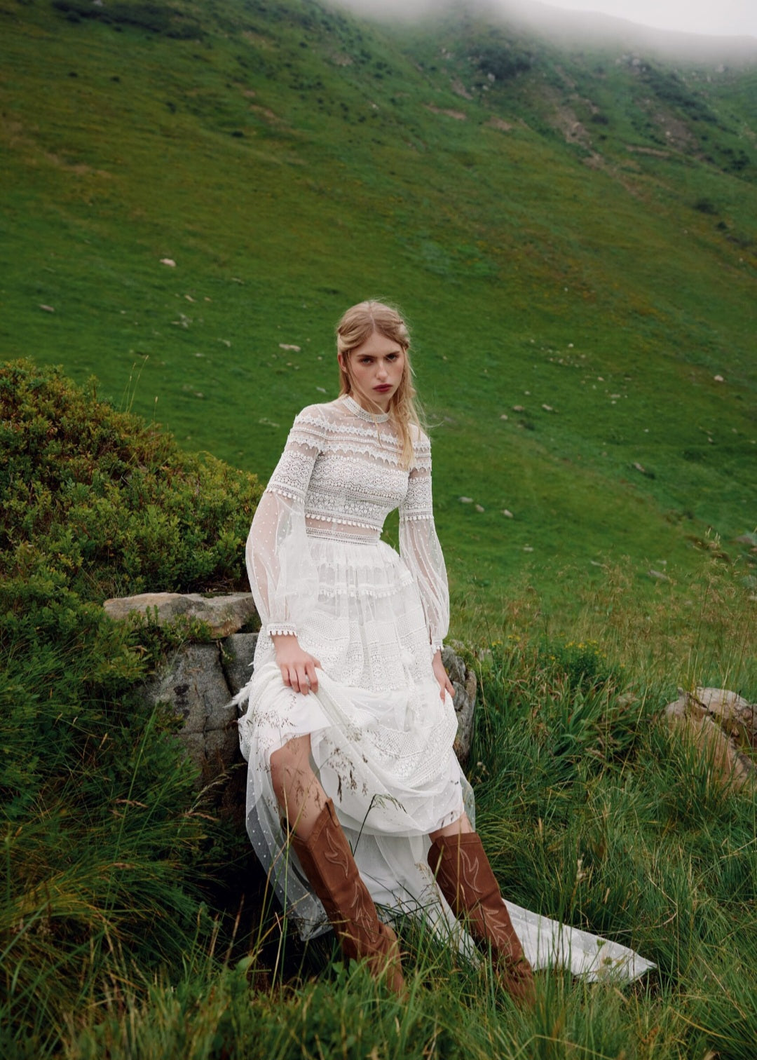 Woman in a white dress and brown boots sitting on a rock in a green, hilly landscape.