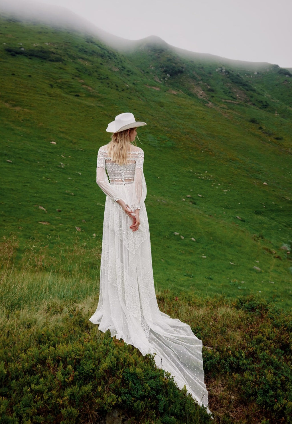 Woman in a white dress and hat standing on a grassy hill with a misty mountain background