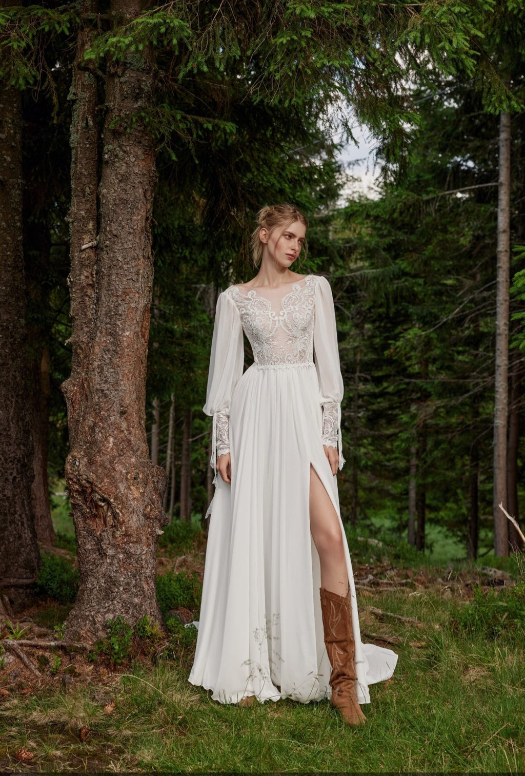 Woman in a white lace dress with long sleeves and a high slit standing in a forest.