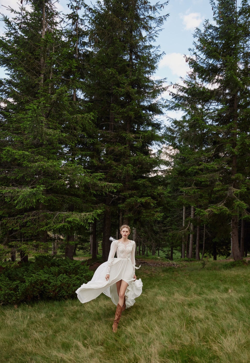 Woman in a white dress standing in a forest with tall trees and green grass.