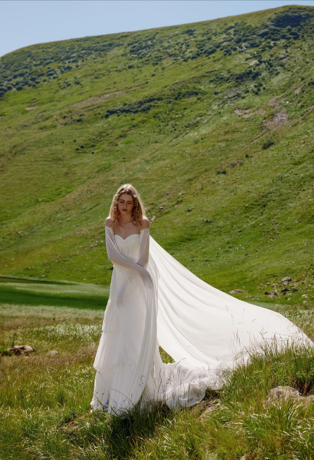 Woman in a white wedding dress standing in a grassy field with mountains in the background