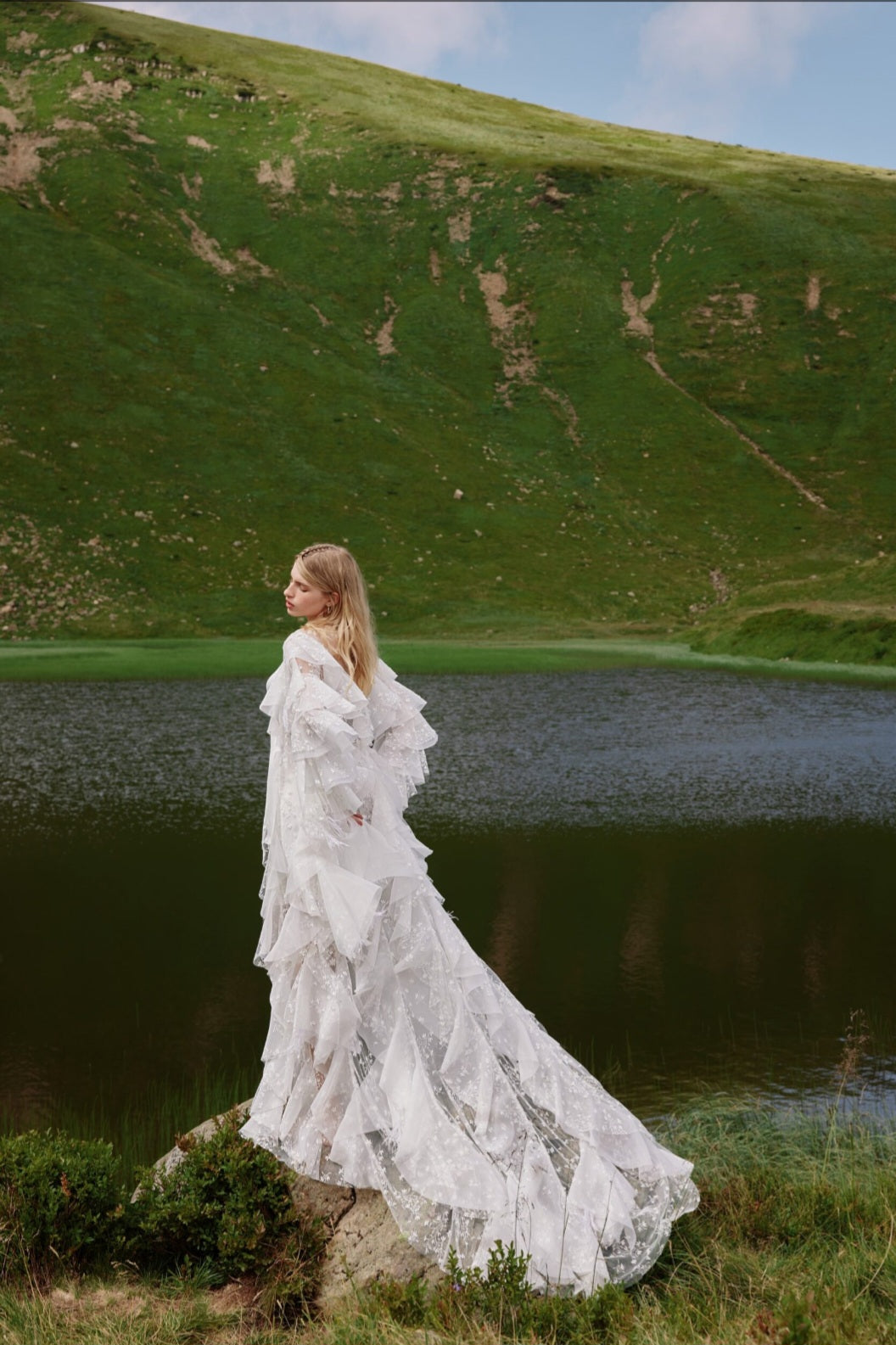 Woman in a white dress standing by a lake with green hills in the background