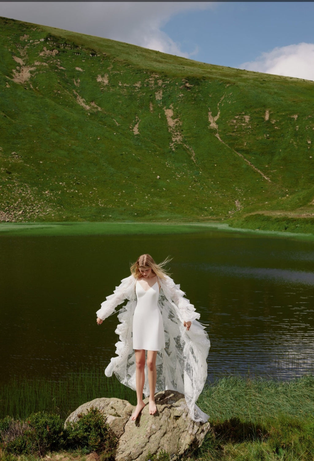 Woman in a white dress standing on a rock by a lake with a green hill in the background