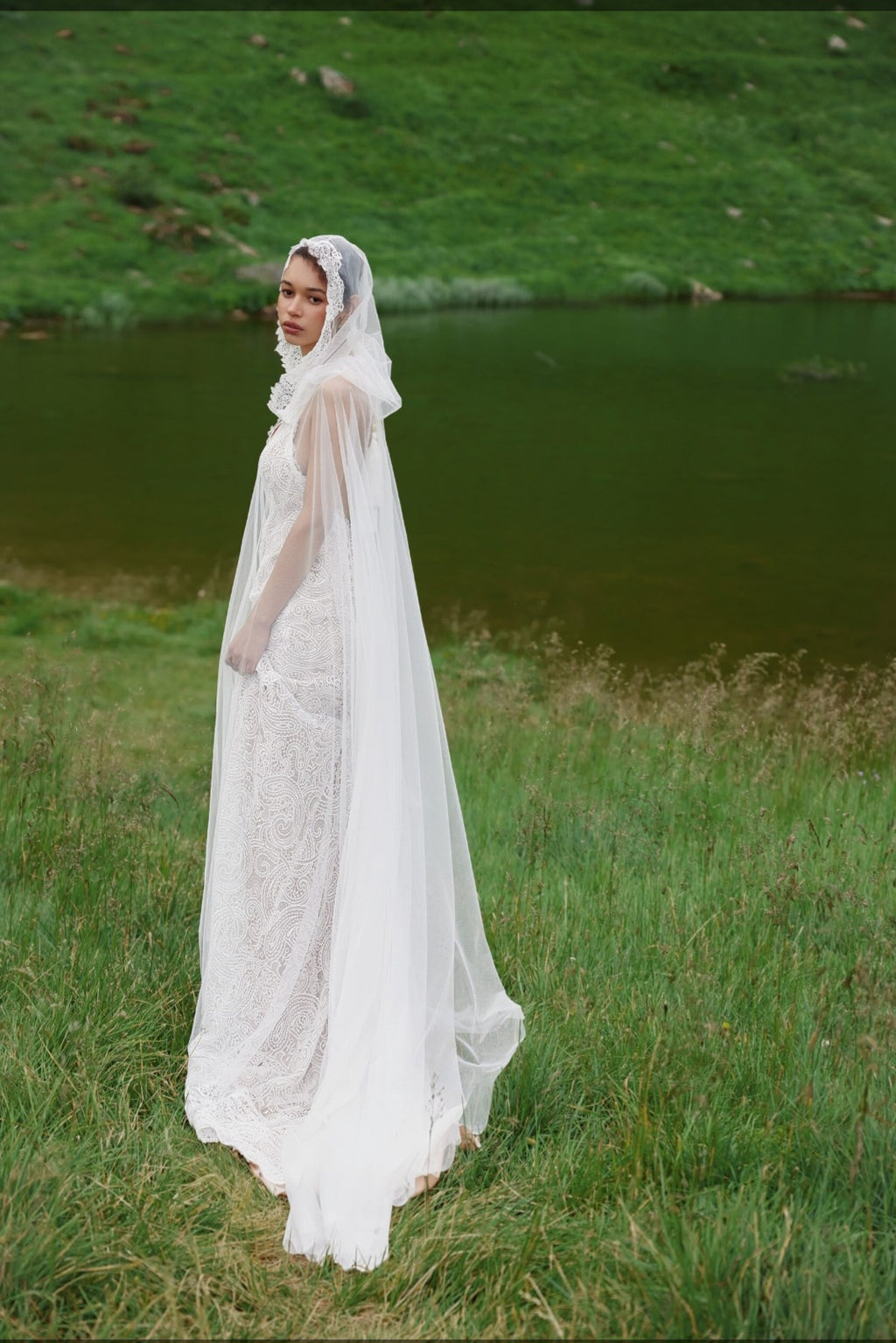 Woman in a white lace dress standing in a grassy field with a body of water in the background