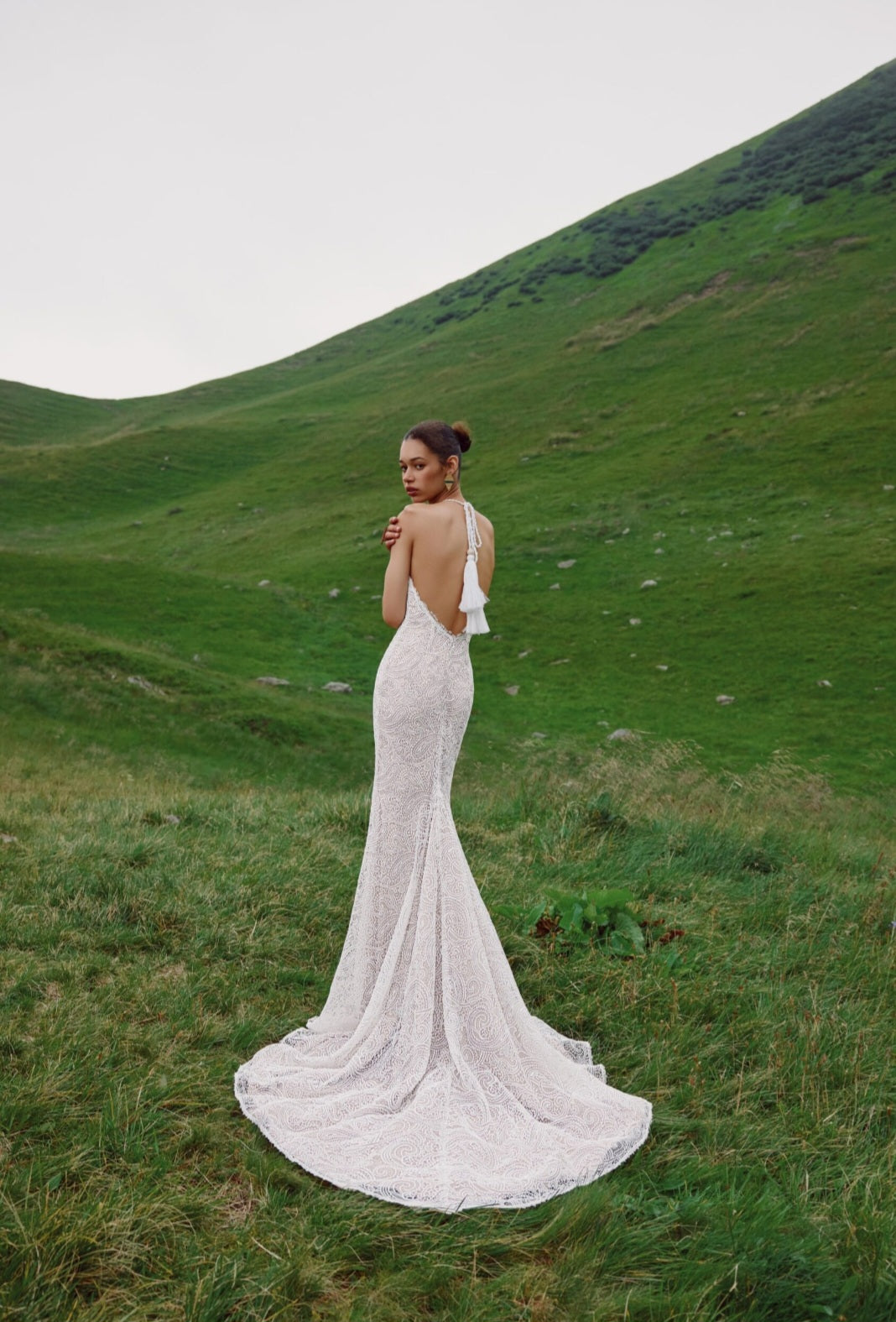 Woman in a white wedding dress standing in a grassy field with hills in the background