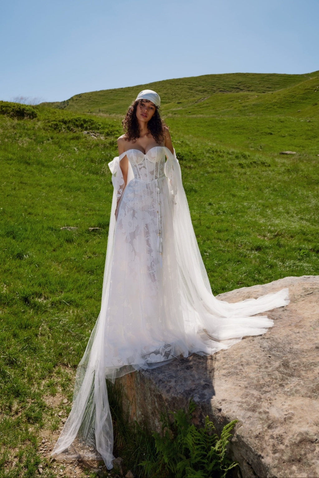 Woman in a white wedding dress standing on a grassy hill with a clear blue sky.