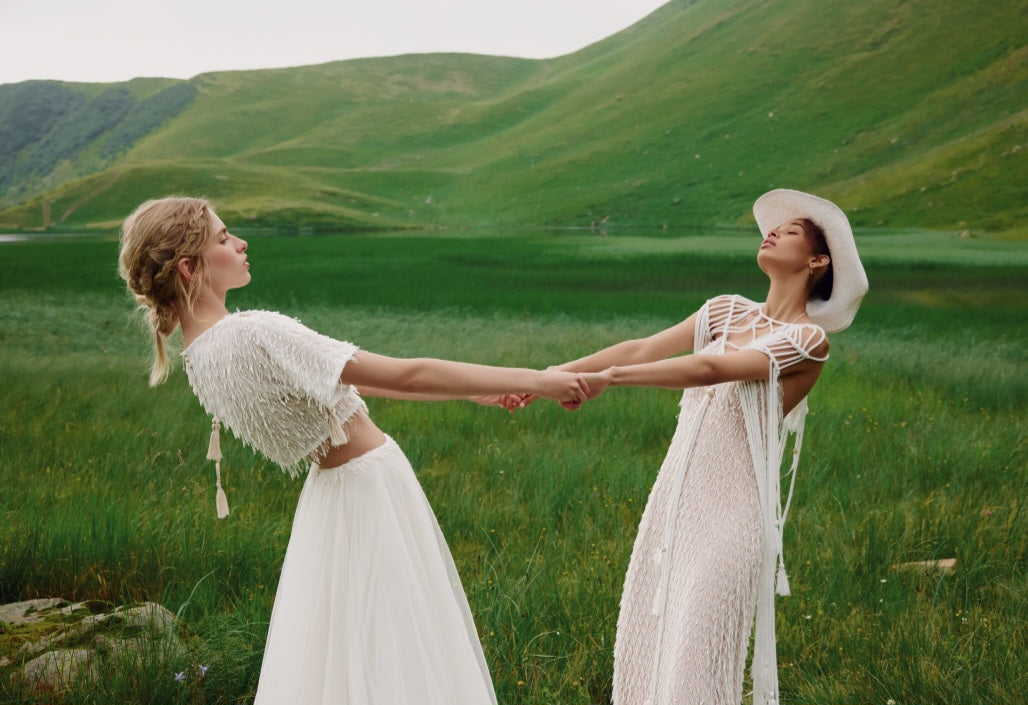 Two women in white dresses holding hands in a green field with rolling hills.