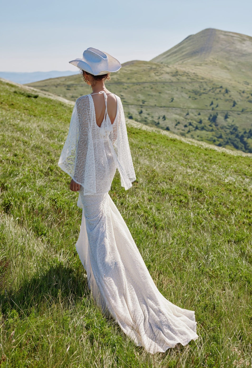 Person in a white dress and hat standing on a grassy hill with mountains in the background