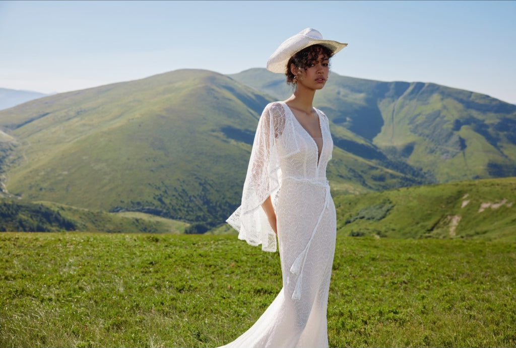 Woman in a white dress and hat standing in a grassy field with mountains in the background