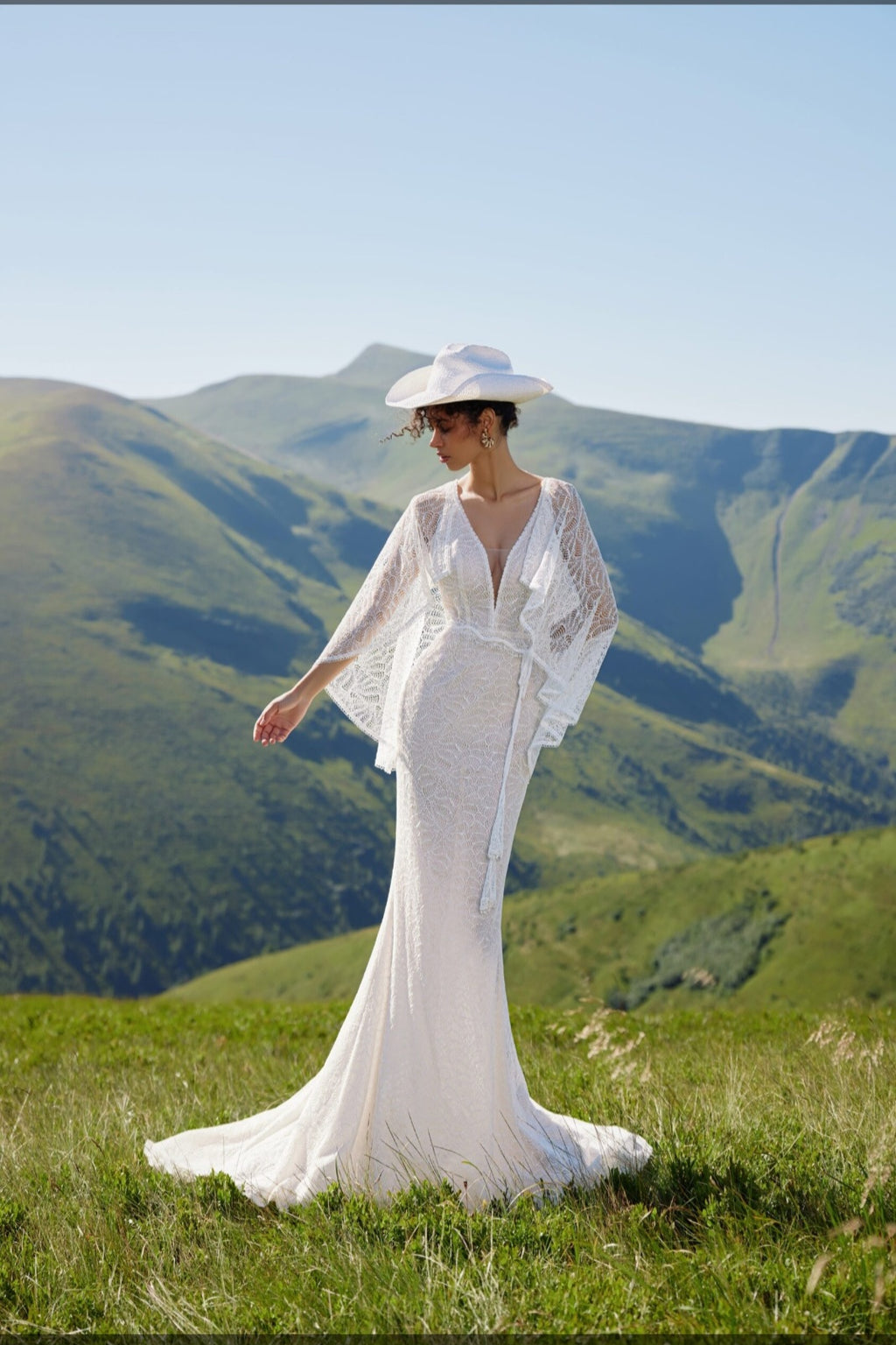 Woman in a white dress and hat standing on a grassy hill with mountains in the background
