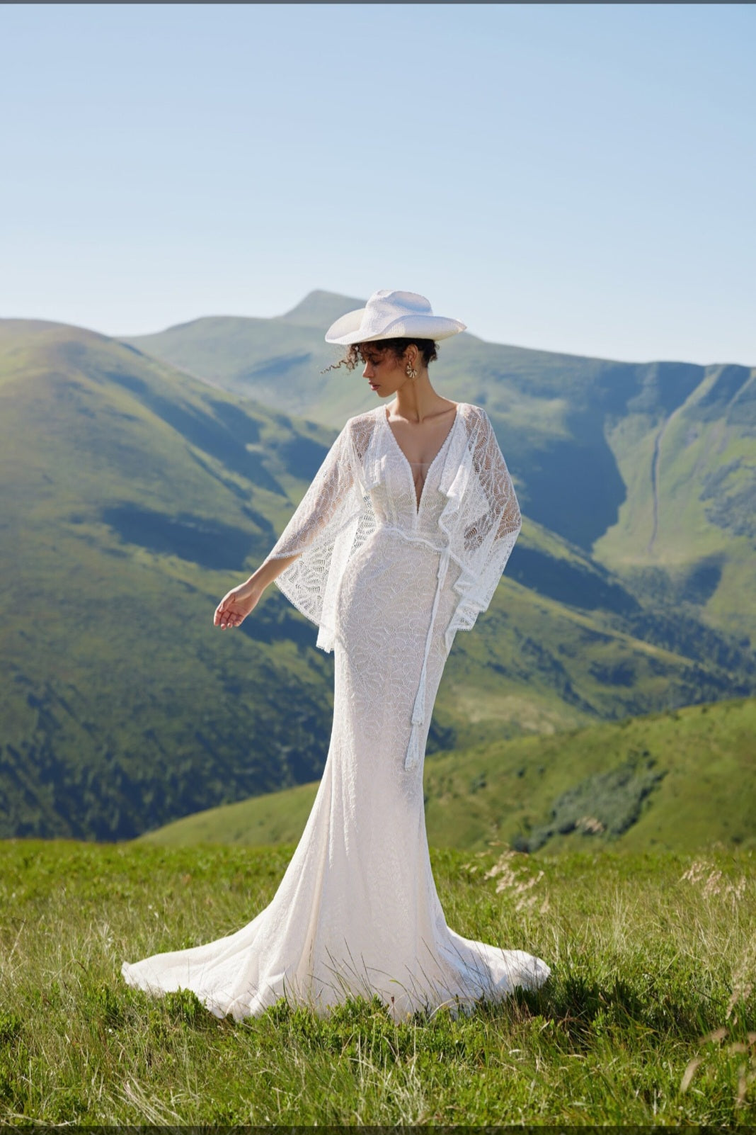 Woman in a white dress and hat standing on a grassy hill with mountains in the background