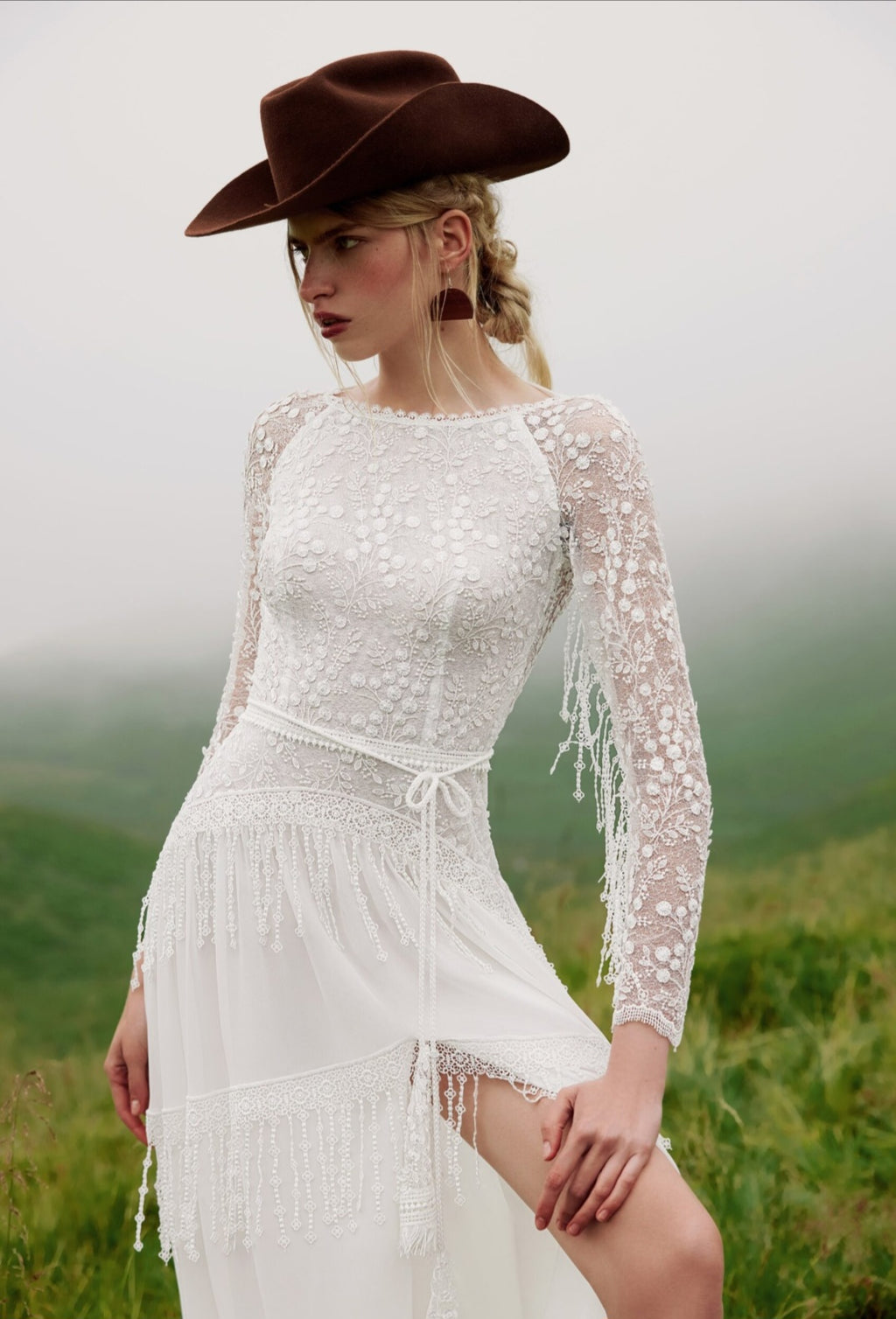 Woman in a white lace dress with fringe and a brown cowboy hat standing in a field.