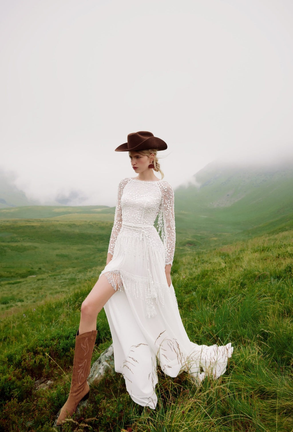 Woman in a white dress and brown boots standing in a grassy field with mountains in the background