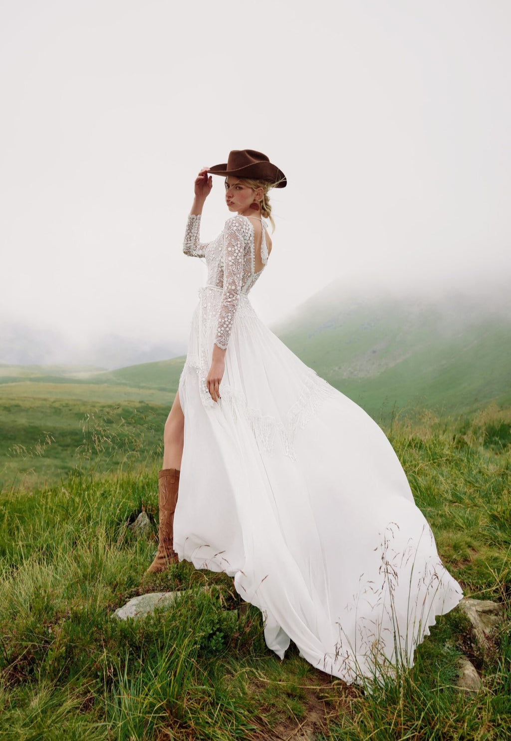 Woman in a white lace dress and brown hat standing in a grassy field with mountains in the background.