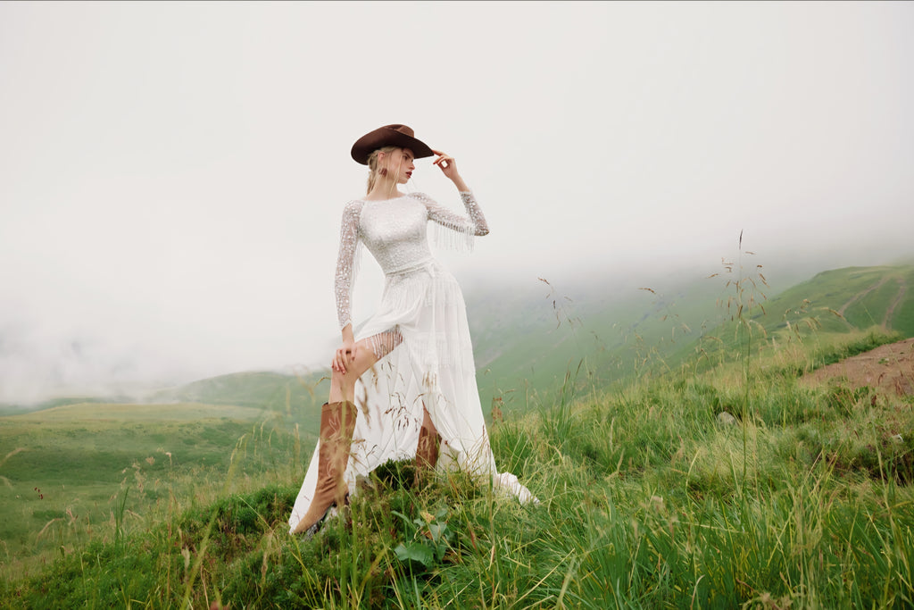 Woman in a white dress and brown hat standing on a grassy hill with foggy mountains in the background