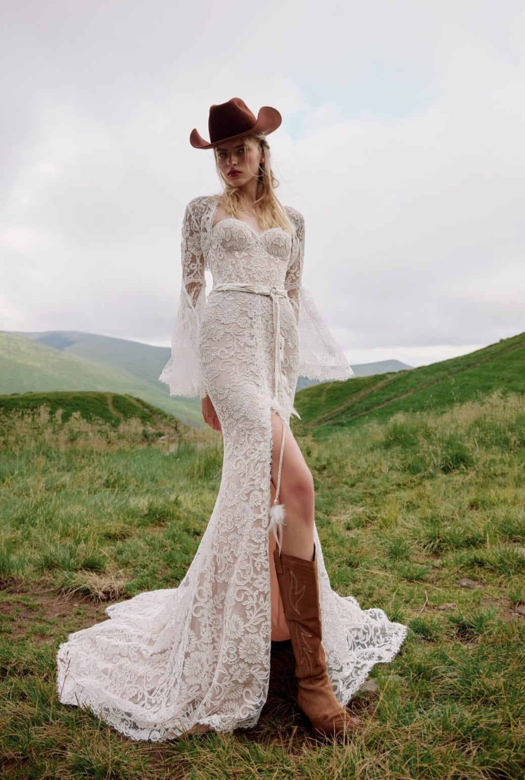 Woman in a lace wedding dress and cowboy hat standing in a grassy field with mountains in the background.