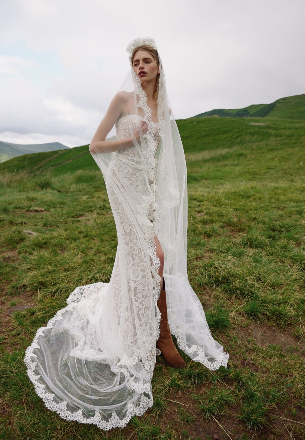 Woman in a white lace wedding dress standing in a grassy field with mountains in the background