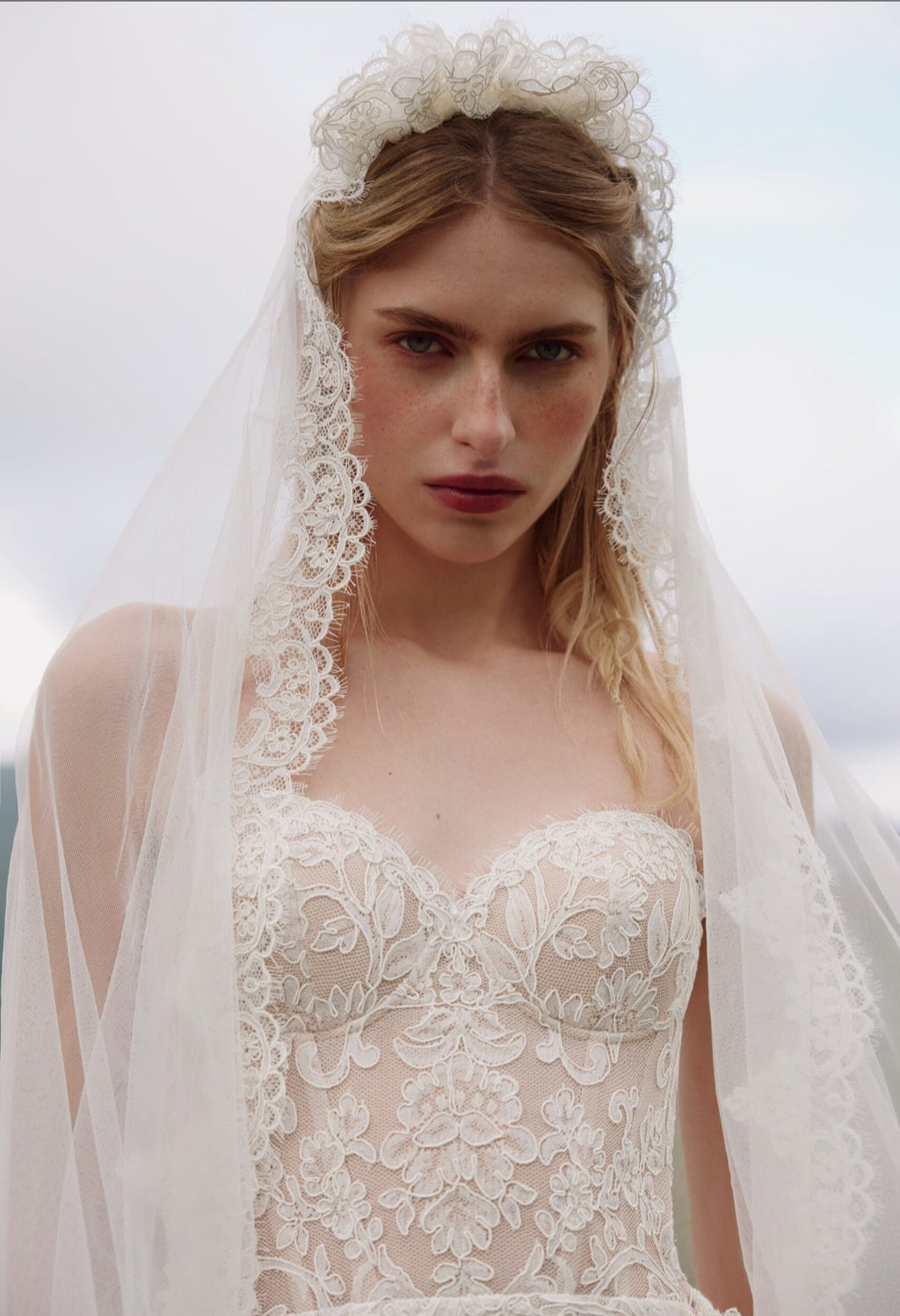 Woman wearing a lace wedding dress and veil against a cloudy sky.
